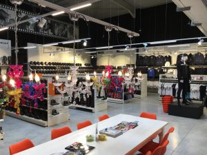 Winkelinterieur met witte tafels, oranje stoelen, verlichte stellingen met schoenen en kleding, kristallen hangende decoraties en feestelijke slingers.