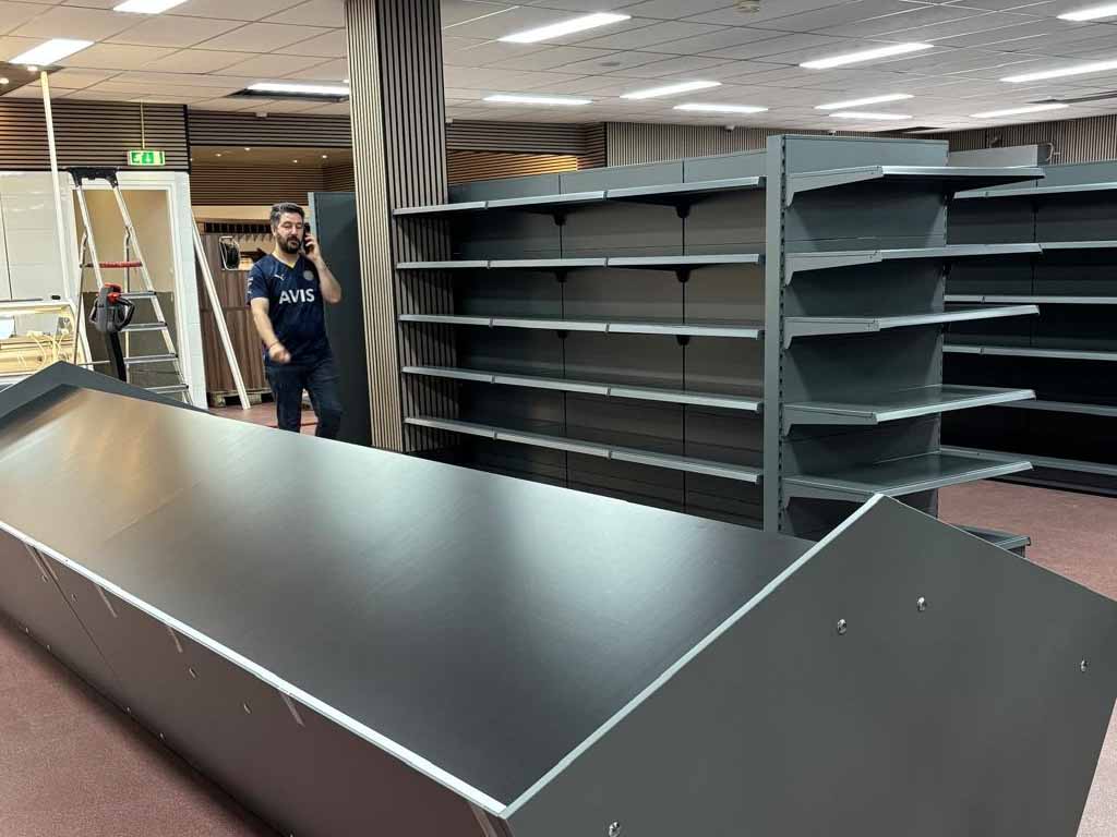 Een man in een donkerblauw shirt staat in een lege winkel met metalen stellingen, een zilveren balie en fluorescente verlichting.