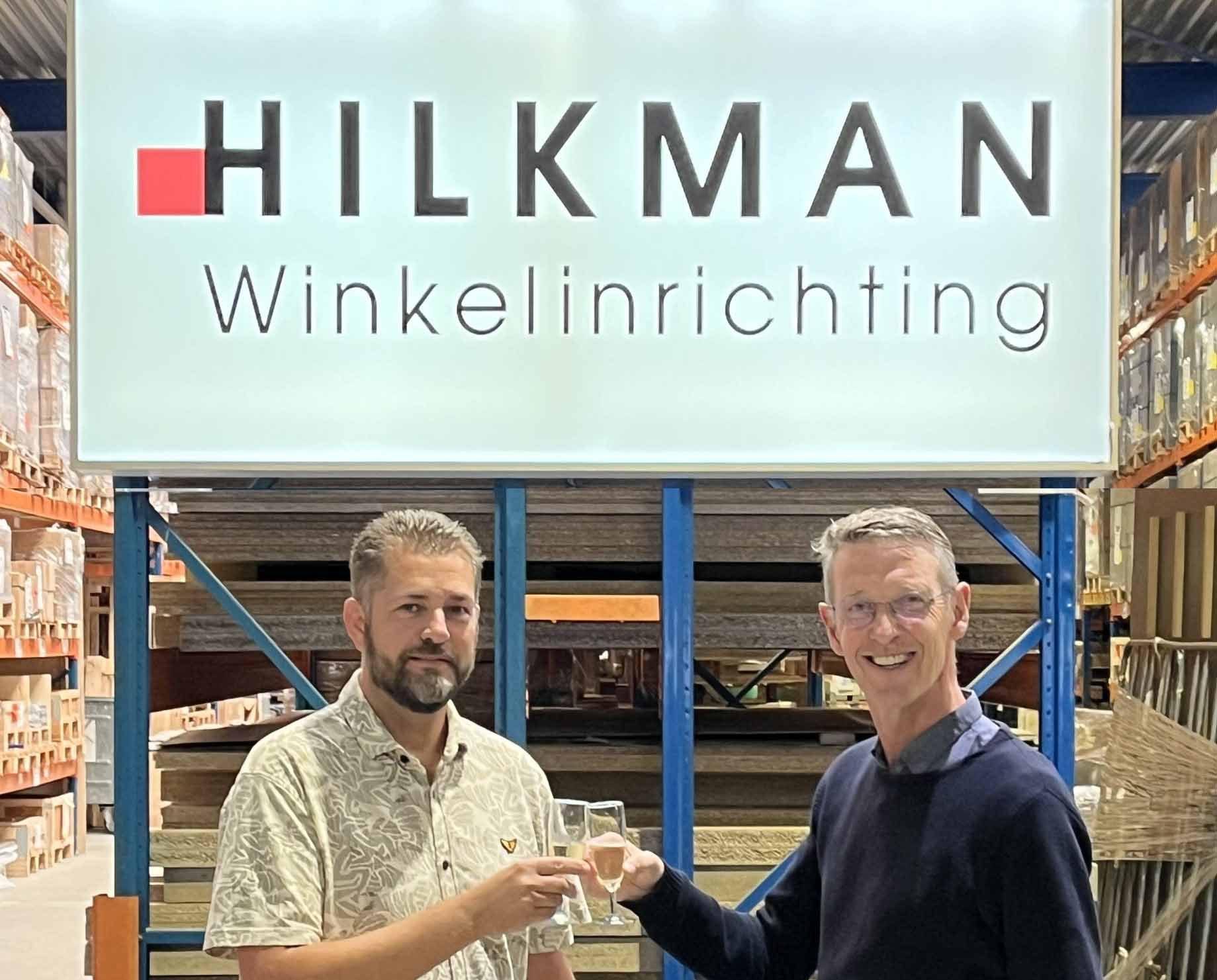 Twee mannen proosten met champagne onder een bord met 'Hilkman Winkelinrichting' in een showroom met stellingen en display.