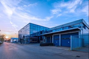 Modern industrieel gebouw met glazen gevel, blauwe deuren en metalen constructie aan een verlaten straat bij zonsondergang.