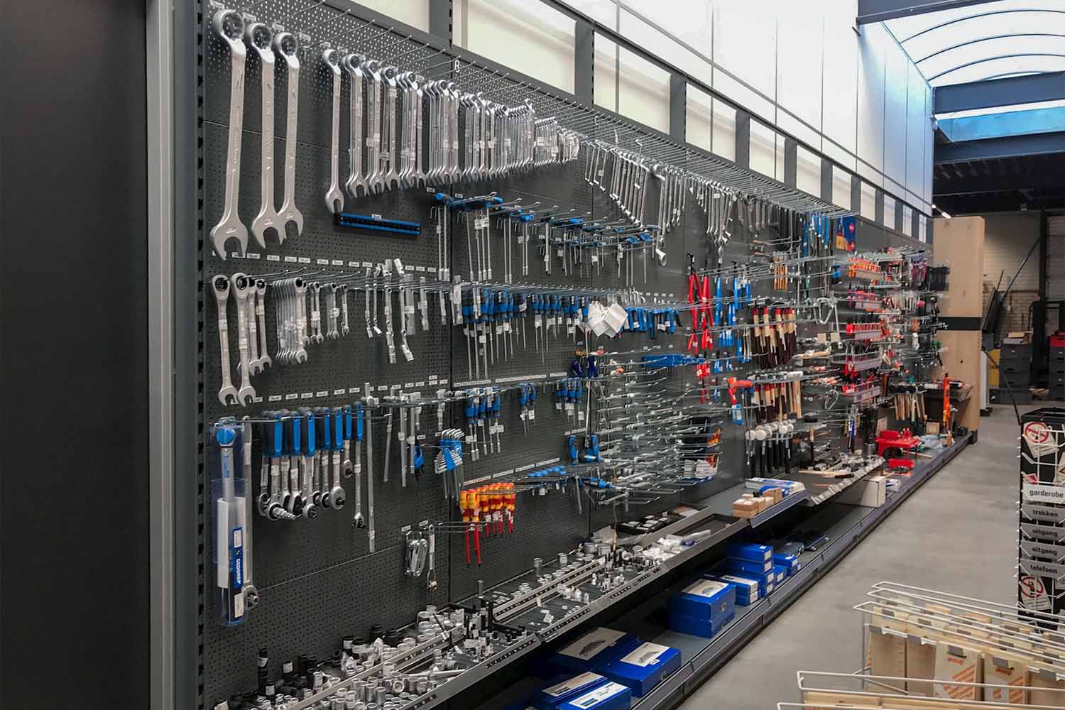 Uitstalling van gereedschap op zwarte pegelborden in een winkel met metalen stellingen, gereedschapskisten en diverse hand- en elektrowerktuigen.