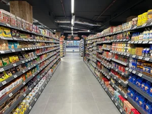 Overzicht van een supermarktgang met zwarte metalen stellingen vol verpakte voedselproducten aan beide zijden en een verharde vloer.