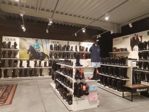 Een moderne laarzenwinkel met witte wandkasten vol bruine en zwarte laarzen, centrale displaytafels en spotverlichting aan het plafond.