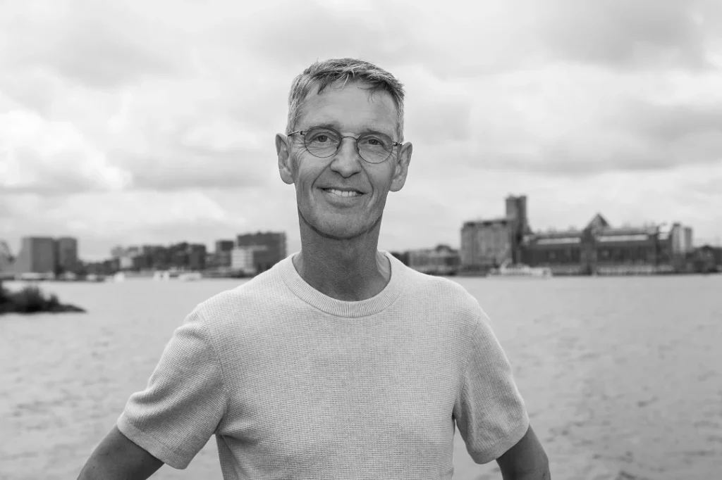Zwart-witfoto van een man met bril en licht shirt die glimlachend voor een waterfront met stadsgebouwen staat.