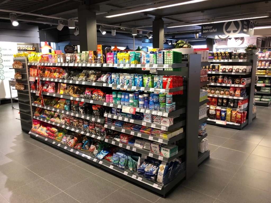 Overzicht van een supermarkt met zwarte metalen schappen vol gekleurde voedselverpakkingen, dranken en snacks onder felle verlichting.
