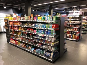 Overzicht van een supermarkt met zwarte metalen schappen vol gekleurde voedselverpakkingen, dranken en snacks onder felle verlichting.