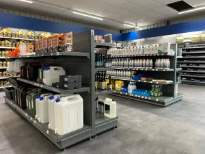 Een moderne winkel met metalen stellingen vol motoroliën, onderhoudsproducten en kannen, tegen blauwe wanden met grijze stellagekasten op de achtergrond.