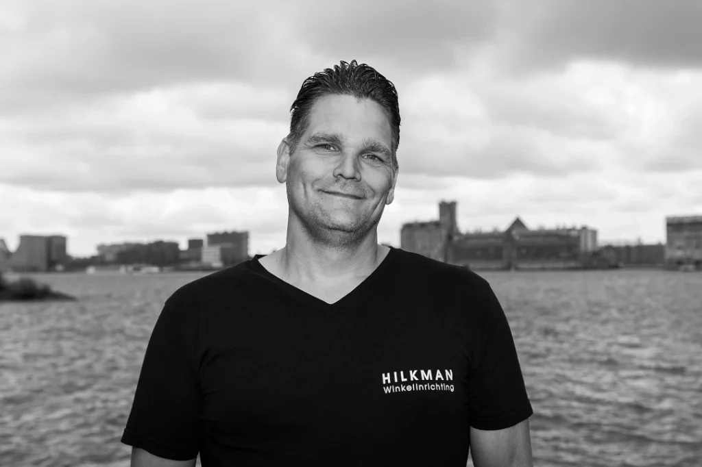 Een man in zwart shirt staat voor een waterspiegel met stadsgezicht op de achtergrond.