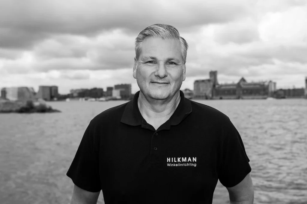 Portretfoto van een man in zwart poloshirt met 'HILKMAN Winkelinrichting' logo, staand voor een waterfront met stadsgebouwen op de achtergrond.