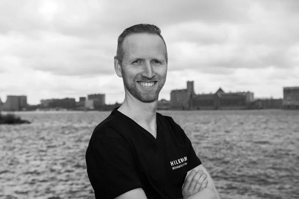 Man in zwart shirt staat glimlachend voor waterkant met stadsskyline op achtergrond.