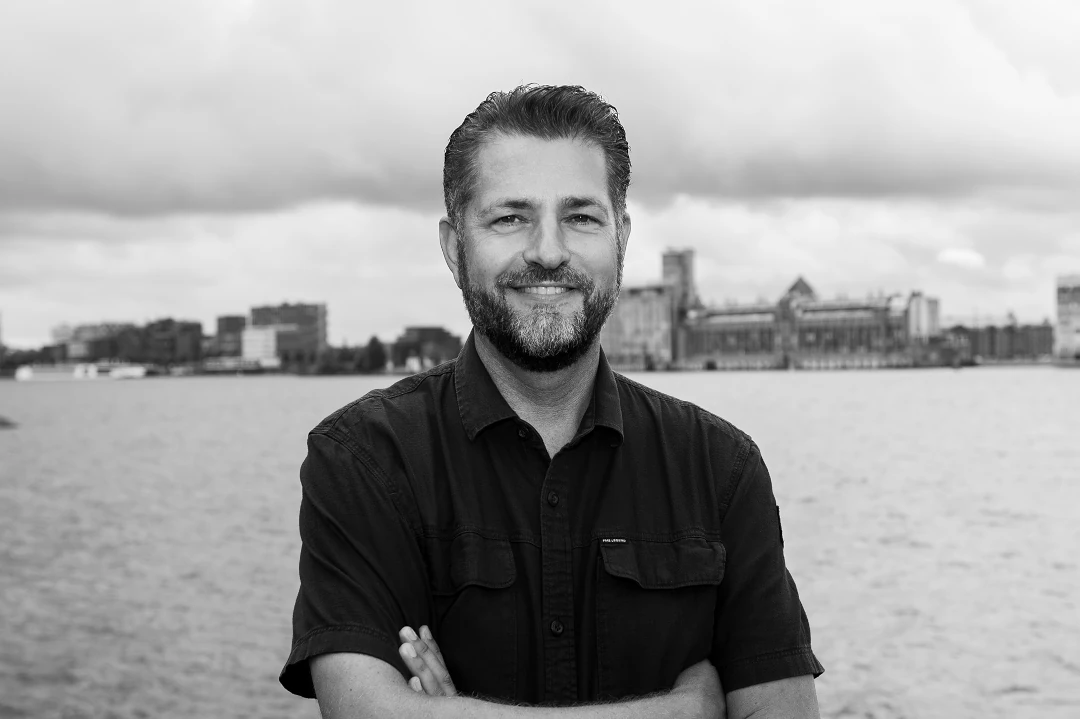 Portretfoto van een man met baard in zwart shirt, staande voor een waterfront met stadsgebouwen op de achtergrond.