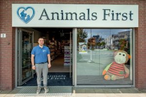 Een medewerker staat voor de ingang van de dierenwinkels Animals First met grote etalageruiten en een knuffeldier in het raam.
