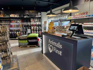 Interieur van een dierenwinkel met toonbank, stellingen met dierenbenodigdheden, snackbar en groen kattenbed op display.
