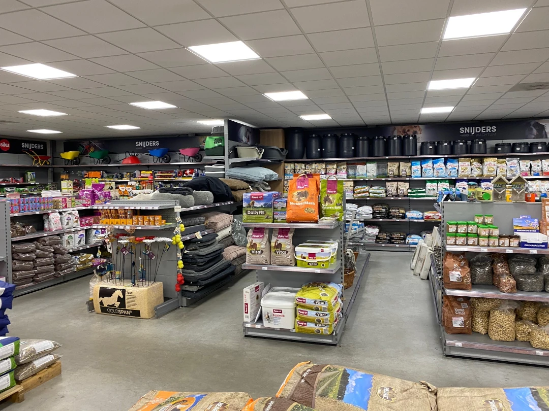 Interieur van een dierenvoerwinkel met metalen stellingen vol voederpakketten, bedden en accessoires onder LED-verlichting.
