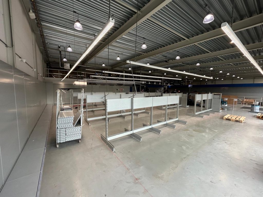 Leeg winkelinterieur met metalen displaystellingen, glazen scheidingswanden en verlichtingsarmaturen onder een golfplaten dak.