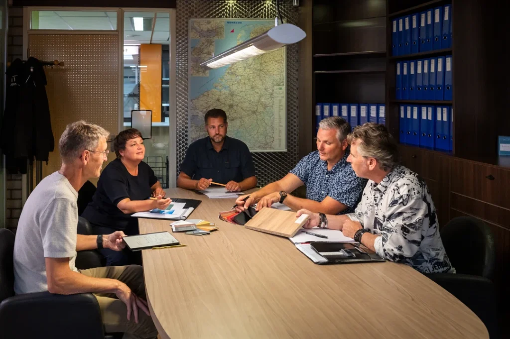 Vijf personen zitten aan een tafel in een moderne kantoorruimte met een kaart aan de muur en blauwe archiefkasten op de achtergrond.