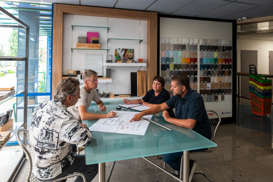 Vier personen bespreken bouwplannen aan een glazen tafel in een winkel met kleurstalen en inrichtingsmateriaal.