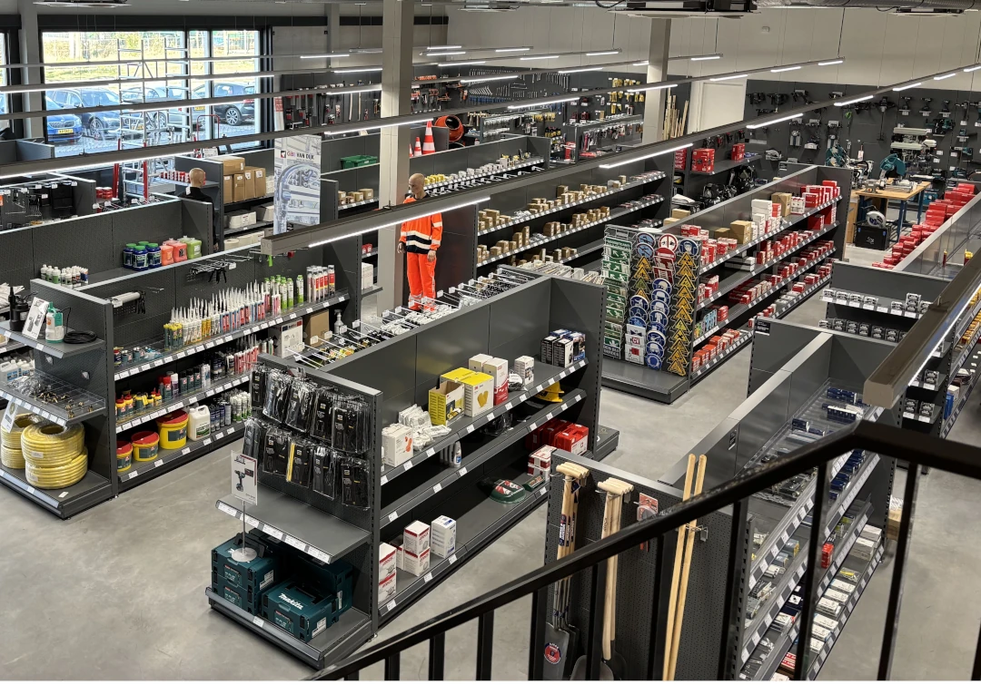 Overzicht van een groot bouwmarkt-winkelinterieur met zwarte metalen stellingen vol gereedschappen, materialen en producten georganiseerd in verschillende secties.