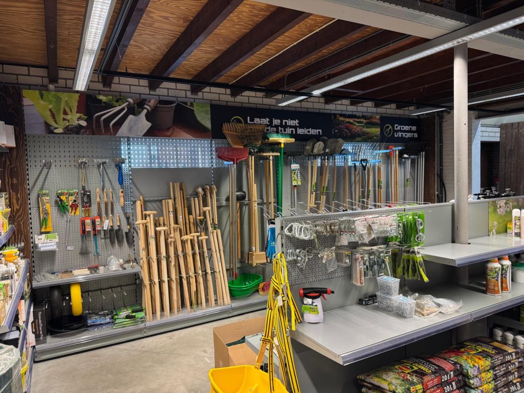 Tuingereedschapwinkel met uitgestalde handgereedschappen, spades en harken op metalen stellingen en pegboards, met verkooptoonbank op de voorgrond.