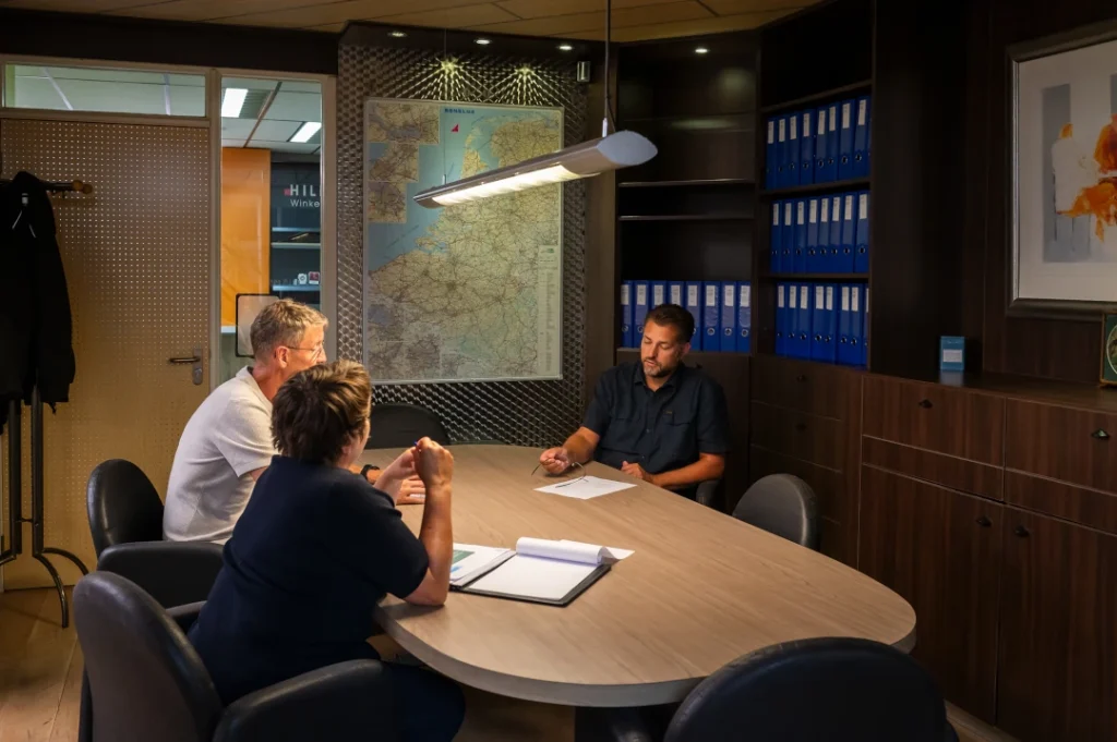 Drie personen zitten aan een houten tafel in een kantoor met kaarten aan de muur, blauwe archiefkasten en donkere houten inbouwkasten.