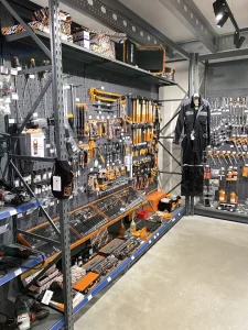 Een gereedschappenwinkel met metalen stellingen vol geel-zwarte gereedschap, displaykasten, een mannequin in werkkleding en diverse bouwmaterialen.