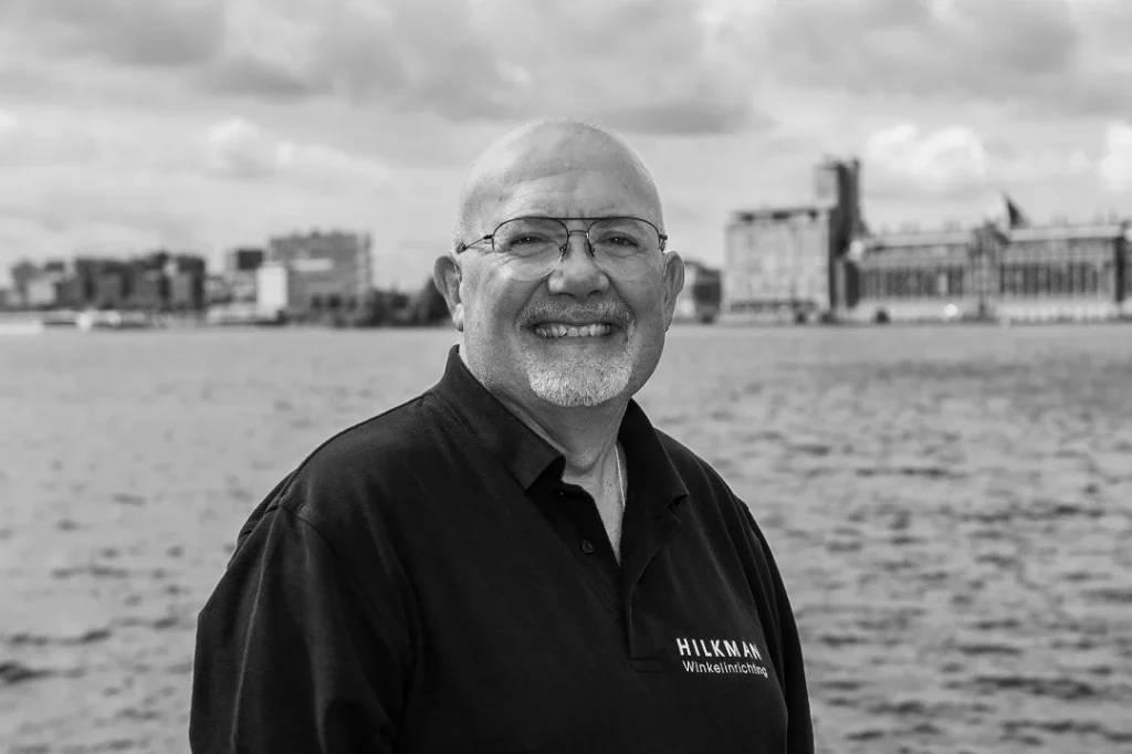 Een man met bril en grijze baard glimlacht naar de camera met een waterfront en stadsgebouwen op de achtergrond.