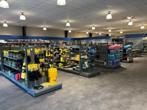 Spacieuze winkel met geel en blauwe metalen stellingen vol gereedschap, autoaccessoires en schoonmaakproducten onder plafondverlichting.