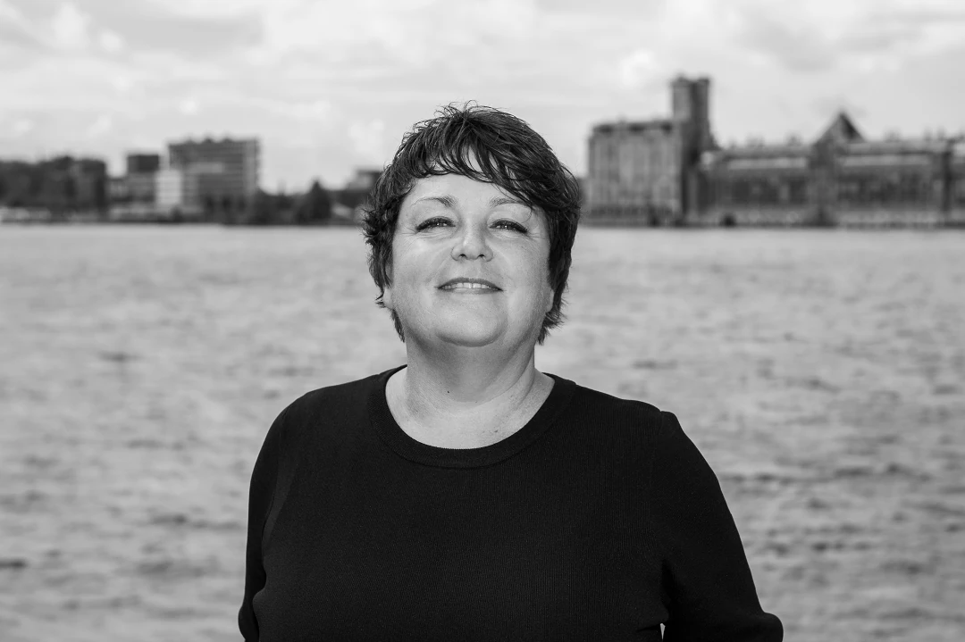 Zwart-witfoto van een vrouw in zwart shirt die naar de camera kijkt, met water en stadsgebouwen op de achtergrond.
