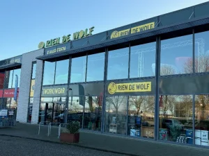Voorgevel van een werkkleding- en uitrustingswinkel met grote glazen ramen en gele letters op donkere gevel langs een parkeerterrein.