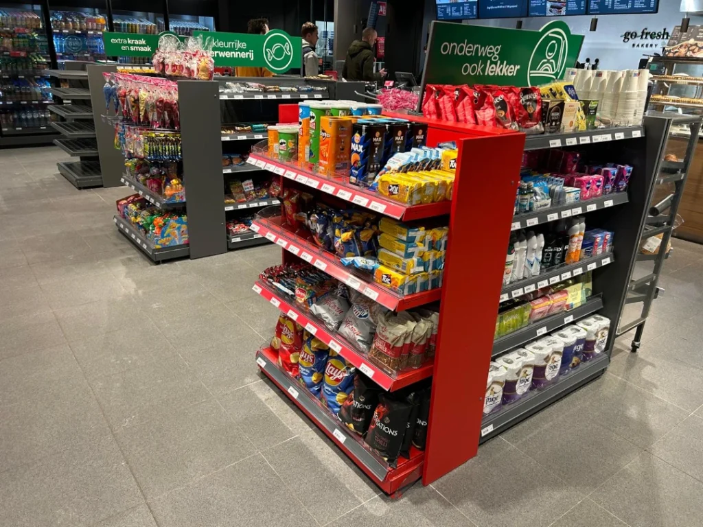 Een rode en zwarte winkelkast met verschillende verdiepingen vol snacks, dranken en snoepgoed in een supermarkt.