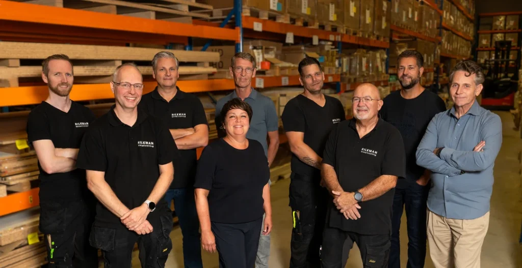 Een team van acht personen staat samen in een magazijn met oranje en blauwe metalen stellingen vol dozen op de achtergrond.