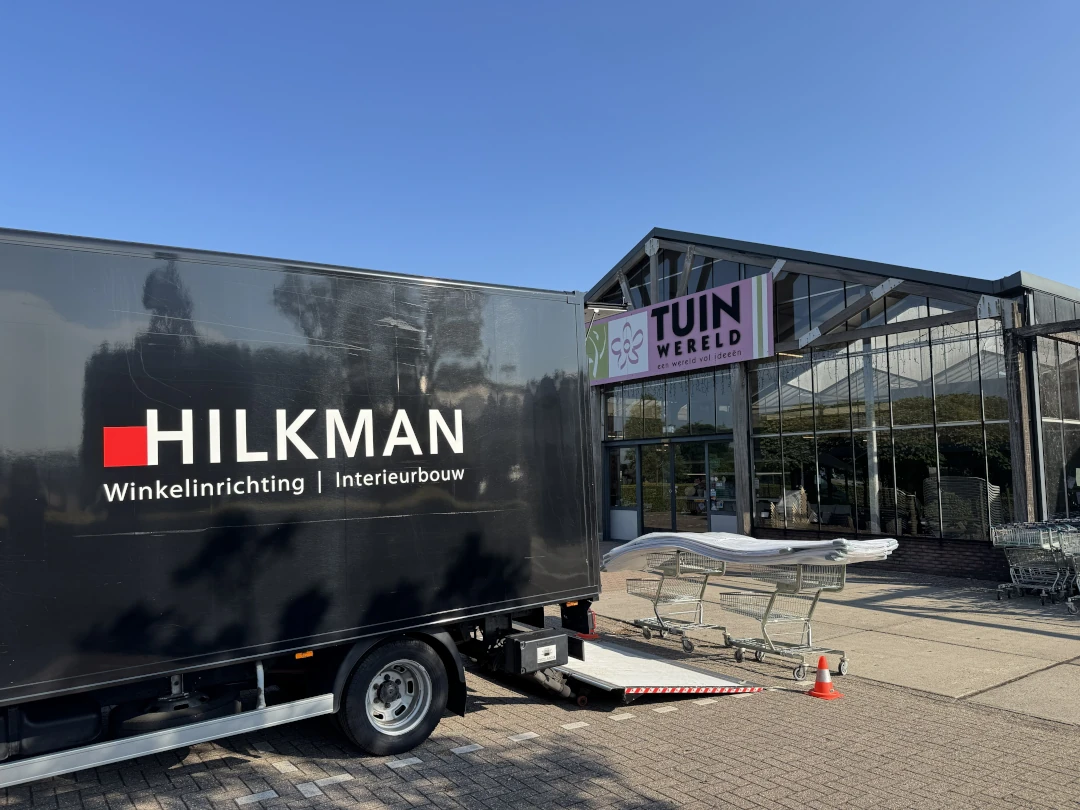 Een zwarte vrachtwagen met winkelinrichting staat voor een glazen showroom met stellingen, winkelwagens en tuinmeubilair op het terrein.