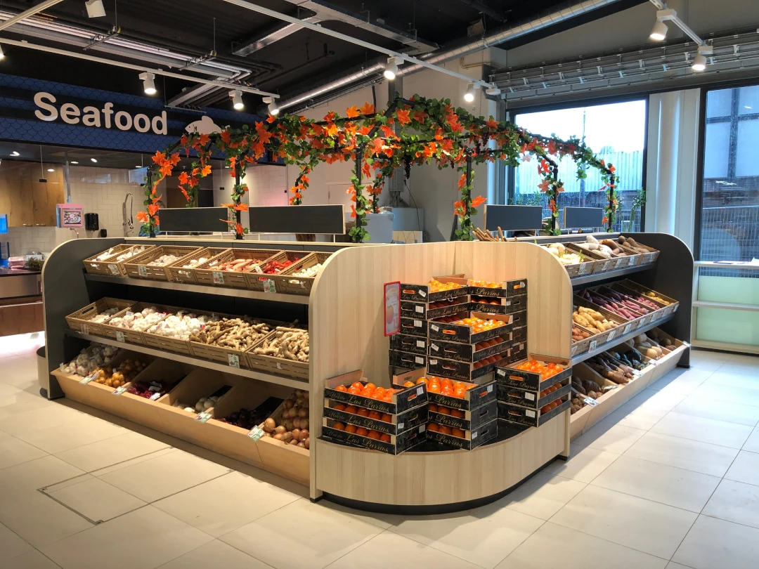 Een moderne supermarktafdeling met houten en zwarte display-balies vol groenten, fruit en andere producten, versierd met oranje bloemenguirlandes.