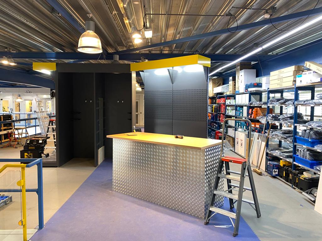 Showroom in een bouwmarkt met gele balie, zwarte displaykasten, metalen stellingen met producten en blauwe vloer.