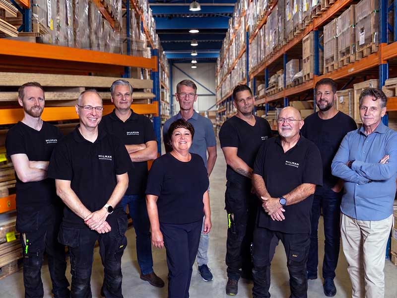 Een team van acht werknemers staat in een groot magazijn met oranje en blauwe metalen stellingen gevuld met dozen en producten.