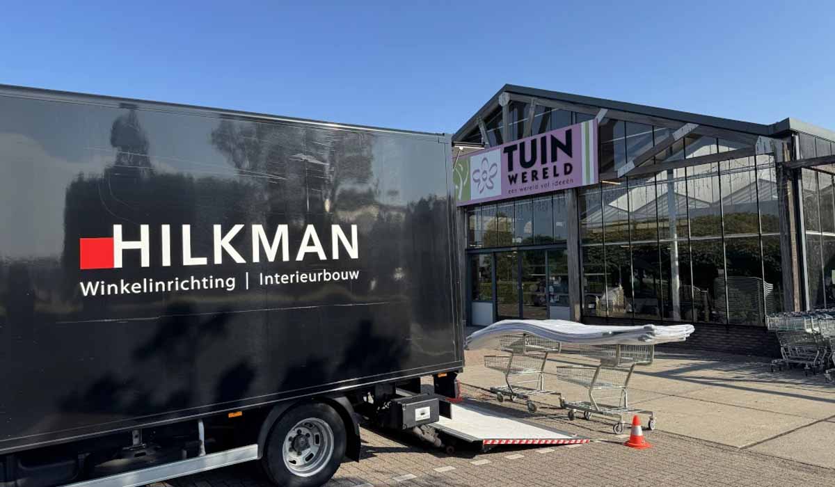 Een zwarte aanhangwagen en een glazen showroomgebouw met winkelinrichting en stellingen op een parkeerterrein onder blauwe hemel.