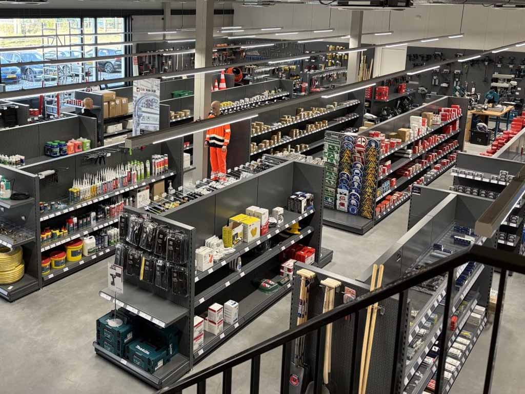Overzicht van een gereedschapwinkel met zwarte metalen stellingen, display-balies en uitgestalde producten zoals verfblikken, boormachines en handgereedschap.