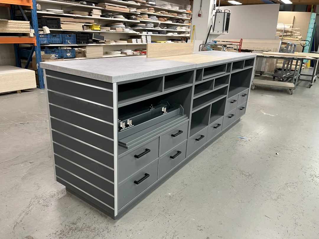 Grijze werkbank met betonnen werkblad, gesloten laden en open vakken in een magazijn met stellingen en gereedschappen.