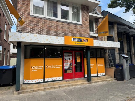 Een geel en oranje winkel aan een straat met grote ramen, rode deur en zwarte stellingen, gelegen naast een bakstenen woonhuis.