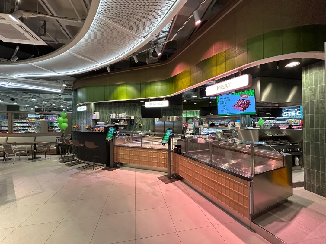 Moderner Supermarkt mit grünen Wandpaneelen, Fleischtheke mit Glaskühlung und digitaler Beschilderung, sowie Kassenbereich mit grünen Dekoballons.