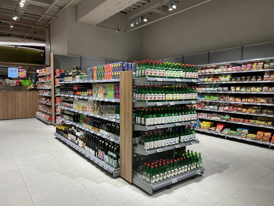Een moderne supermarkt met metalen stellingen gevuld met dranken en voedingsproducten, een centrale display met flessen en een houten balie op de achtergrond.