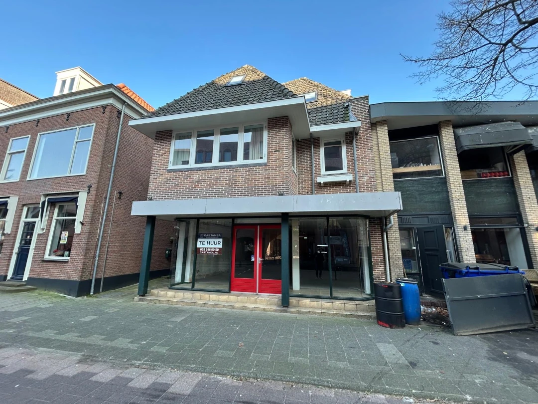 Straatgevel van een winkelgebouw met rode deur, witte luifel en bakstenen voorgevel, naast woningen in een Nederlands dorp.