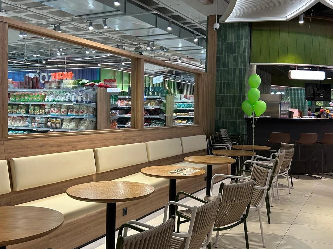 Modern ingerichte winkelruimte met houten zitbank, ronde tafels, groene wandtegels en grote raampartij met productenstellingen.