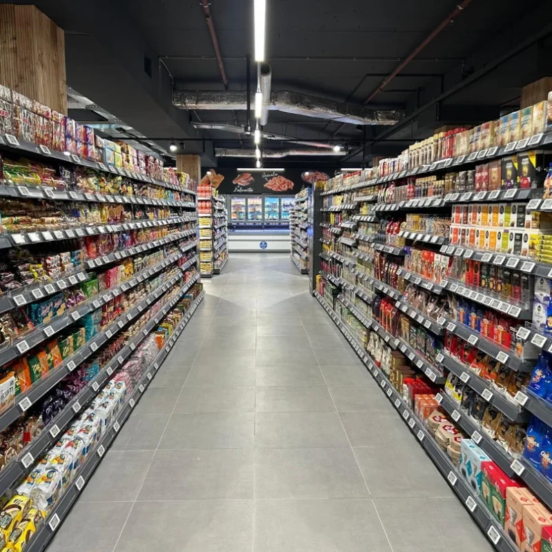 Overzicht van een supermarktgang met zwarte metalen stellingen vol verpakte voedselproducten aan beide zijden en een verharde vloer.