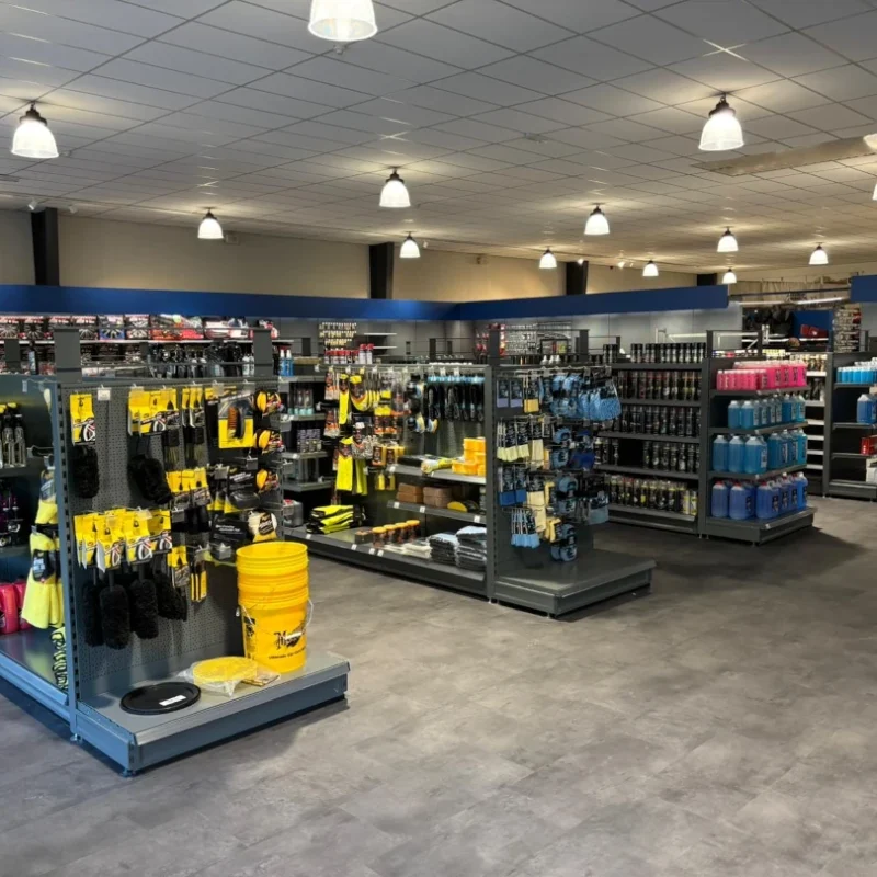 Spacieuze winkel met geel en blauwe metalen stellingen vol gereedschap, autoaccessoires en schoonmaakproducten onder plafondverlichting.