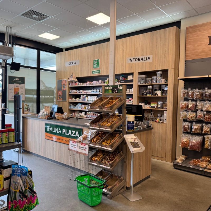 Winkelinterieur van een voedselwinkel met houten wandafwerking, kassabalie, infobalie, metalen stellingen met snacks en droogwaren, en een groene winkelwagen.