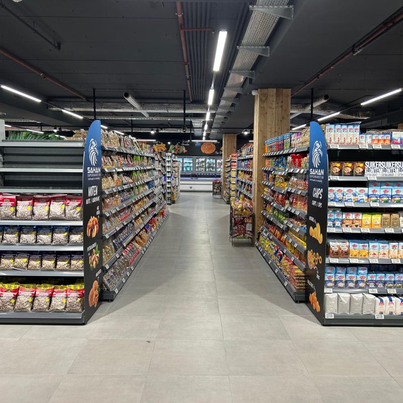 Hypermarkt-gangpad met zwarte metalen stellingen vol verpakte snacks en chips aan beide zijden, onder heldere LED-verlichting.