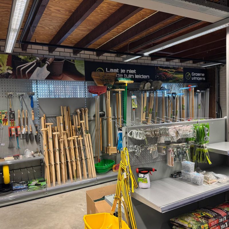 Tuingereedschapwinkel met uitgestalde handgereedschappen, spades en harken op metalen stellingen en pegboards, met verkooptoonbank op de voorgrond.